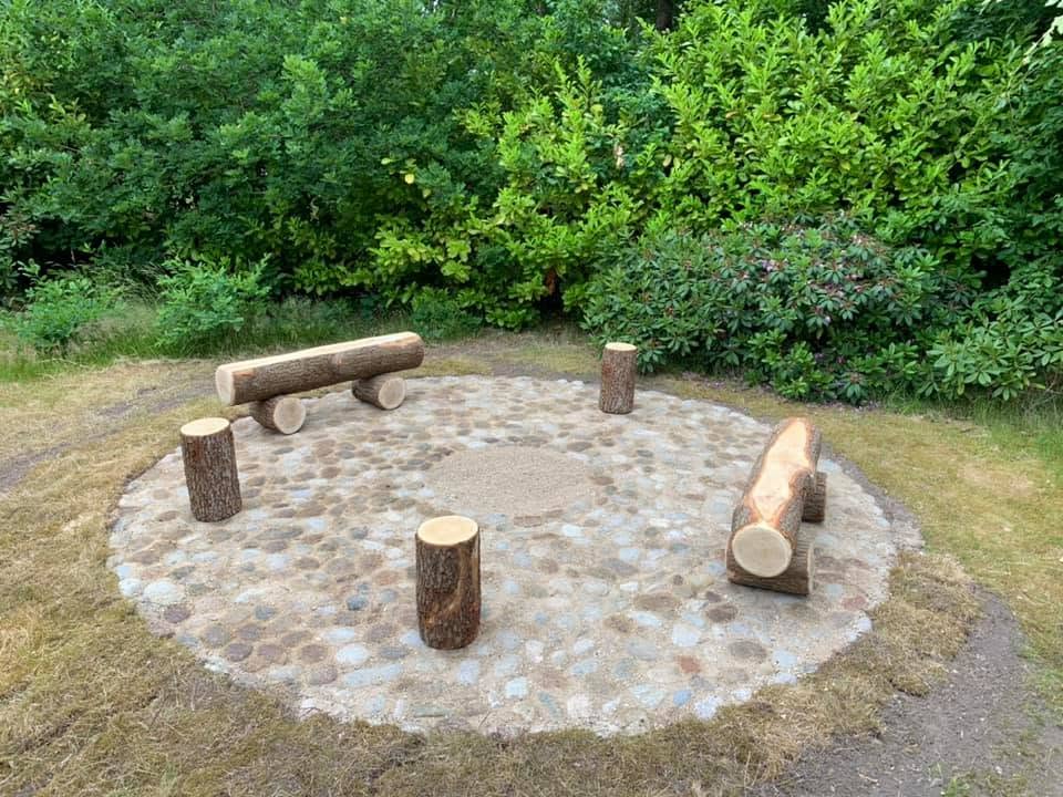 Færdig cirkulær bålplads i marksten med bænke og stubbe omkring sandcirklen